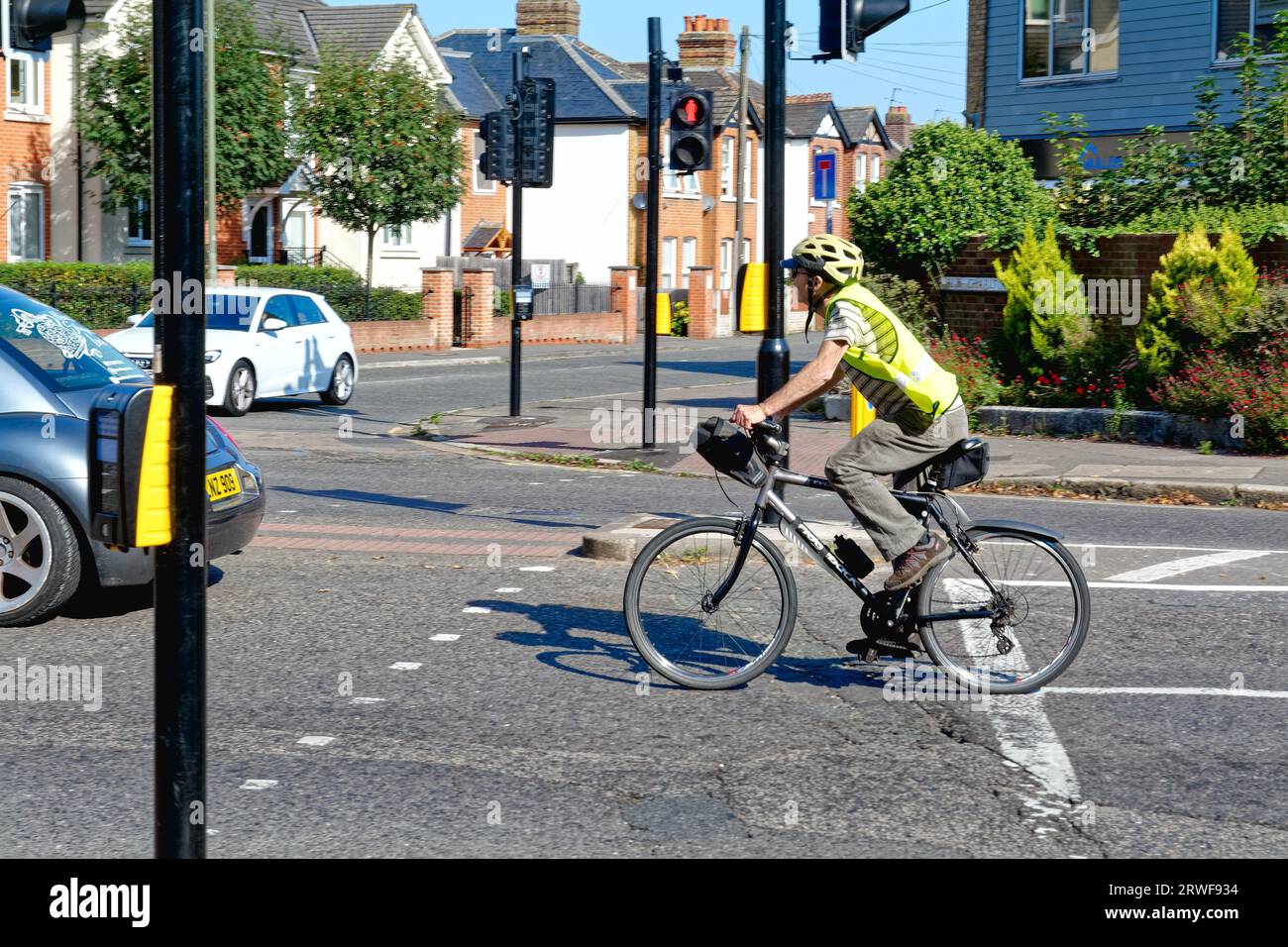 Un anziano ciclista maschile con casco e giacca hi viz che affronta il traffico su un incrocio suburbano, Shepperton Surrey Inghilterra Regno Unito Foto Stock