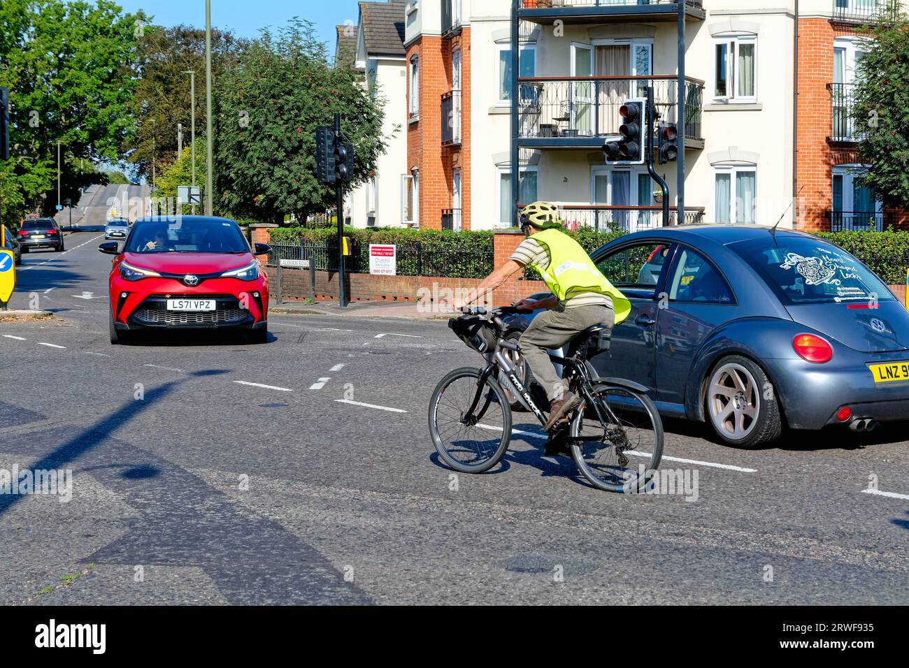 Un anziano ciclista maschile con casco e giacca hi viz che affronta il traffico su un incrocio suburbano, Shepperton Surrey Inghilterra Regno Unito Foto Stock