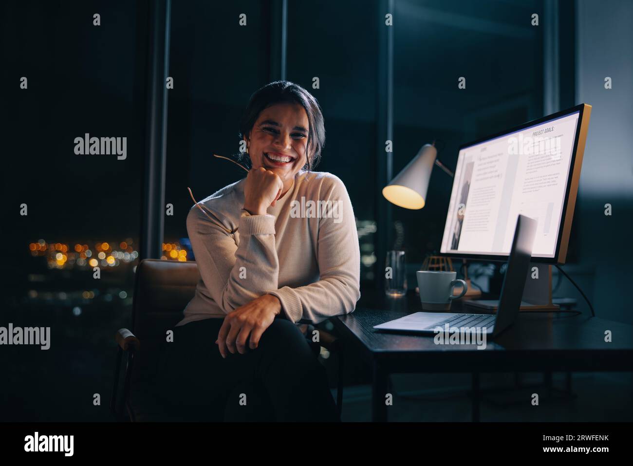 Una donna d'affari felice lavora fino a tardi con il suo computer portatile e il suo computer in un ufficio notturno. Pianifica ed esegue strategie di content marketing al di fuori dell'orario di lavoro, S Foto Stock