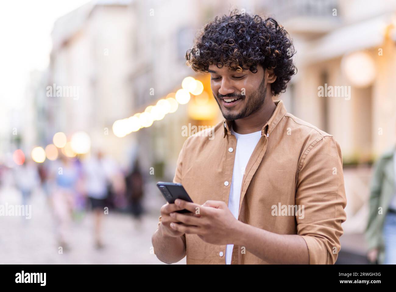 Giovane indiano sorridente che sta fuori e usa il telefono, invia SMS, chatta, chiama. Foto Stock
