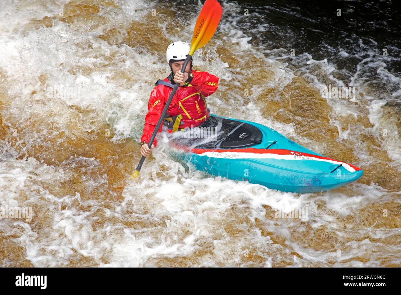 Canoista con giacca rossa e casco in canoa blu che cavalca le rapide in slalom Whitewater nel fiume Tryweryn presso il National Whitewater Centre Foto Stock