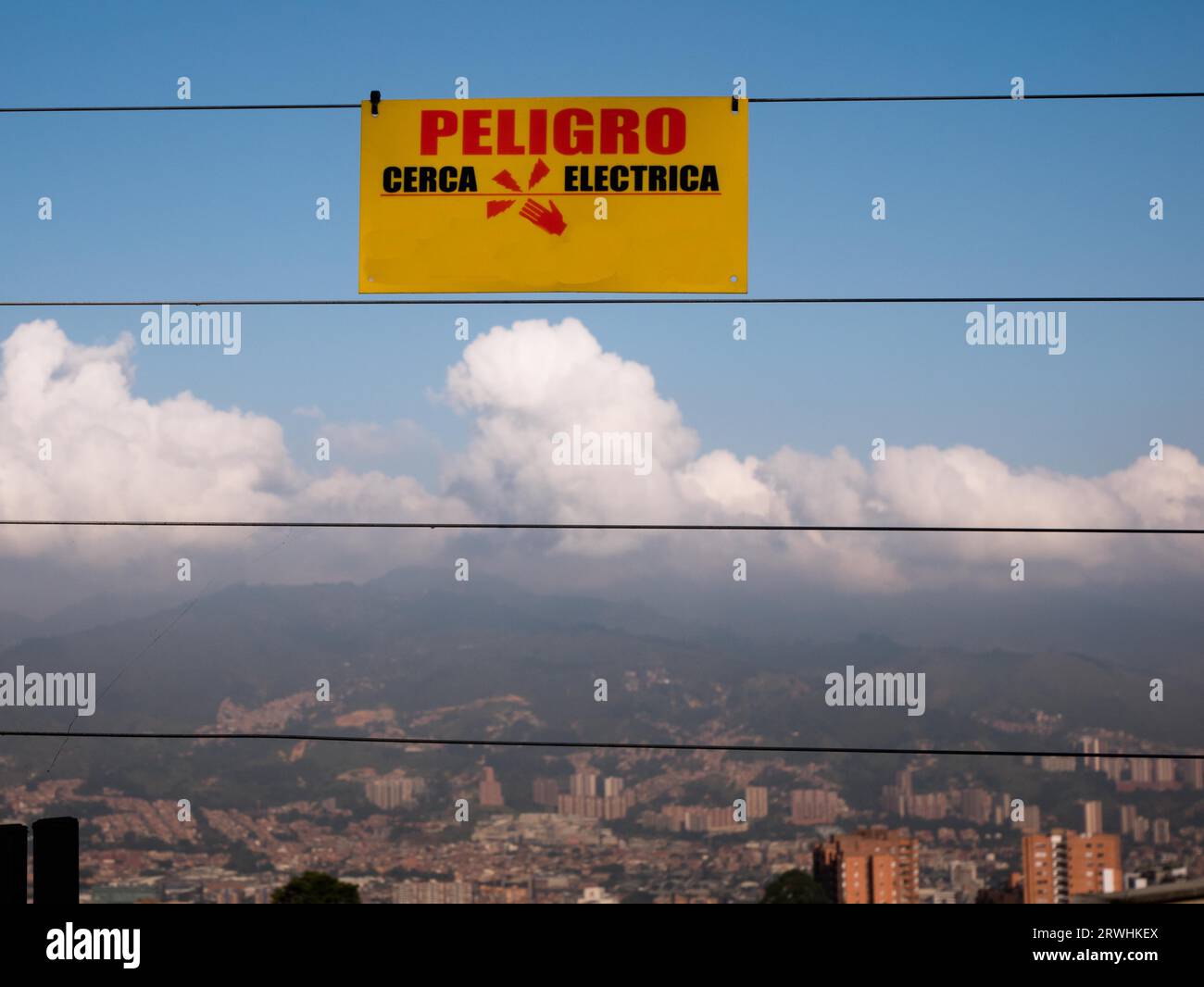 cartello di avvertimento giallo in spagnolo appeso a una recinzione elettrica con lo skyline di una città sullo sfondo Foto Stock