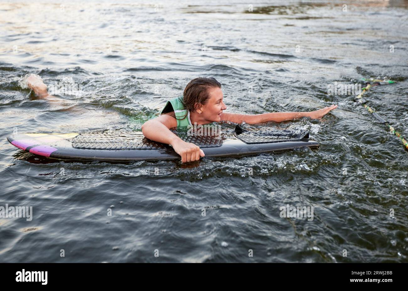 La giovane ragazza sportiva con un giubbotto di salvataggio verde nuota in acqua con un wakeboard in mano. Una sportiva felice si sta preparando per un ripido wakeboa Foto Stock
