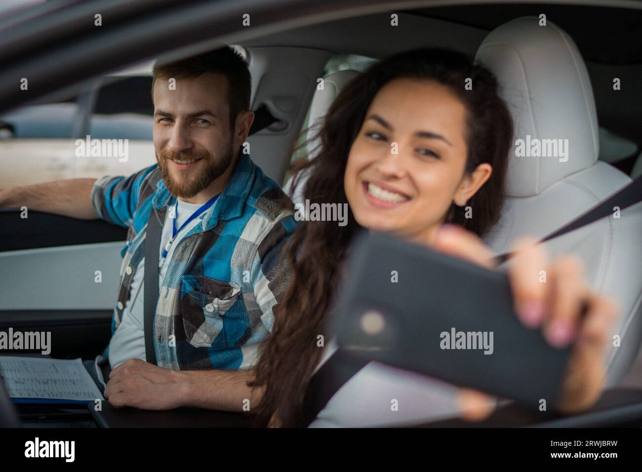 Donna felice che fa selfie e usa lo smartphone, istruttore maschio seduto accanto a lei in auto. Test di guida, corsi per conducenti, concetto di esame Foto Stock