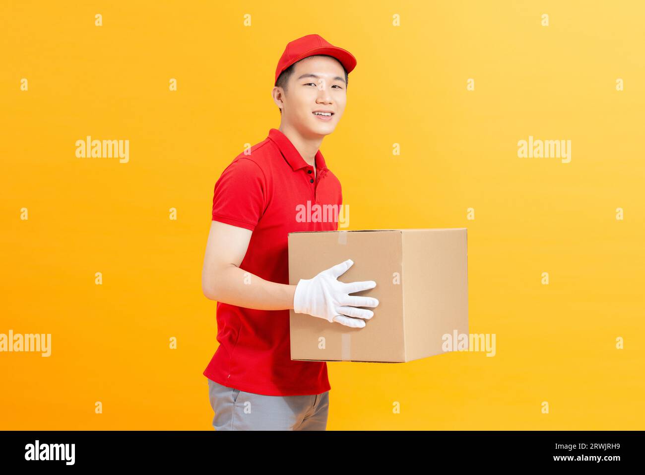 Un giovane e felice addetto alla consegna con tappo rosso in piedi con una cassetta postale isolata su sfondo giallo. Foto Stock