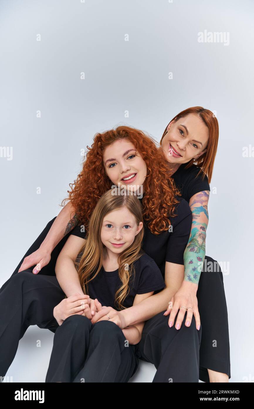 famiglia felice con i capelli rossi seduti in abiti coordinati su sfondo grigio, tre generazioni Foto Stock