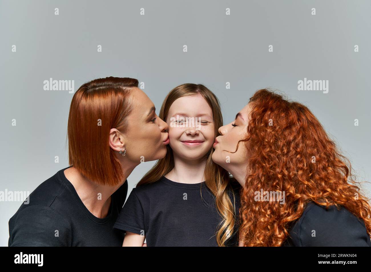 concetto di generazione femminile, donne rosse che baciano guance sorridenti di ragazza su sfondo grigio Foto Stock