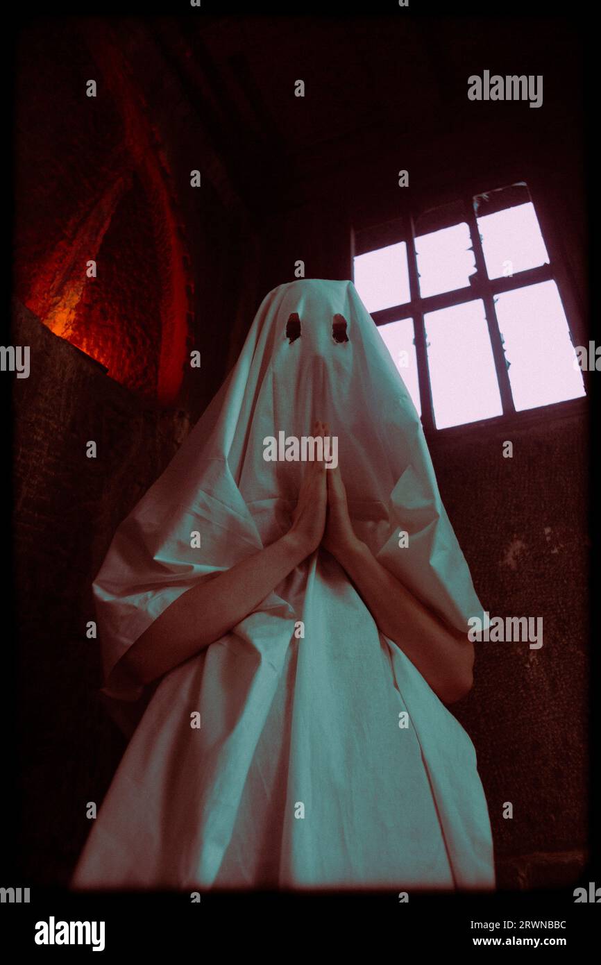 Una donna vestita con un abito bianco spettrale con buchi per gli occhi si trova in una cripta, nel profondo della preghiera. L'occultismo, le sette e la religione. Il costume della donna Foto Stock