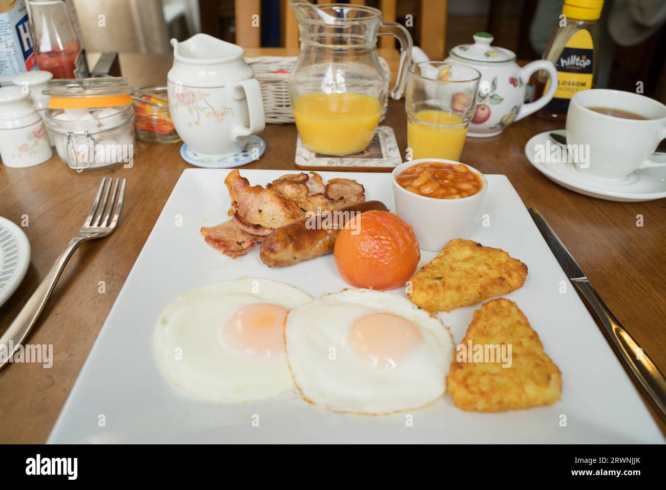 Colazione all'inglese completa con un piatto, pancetta, salsiccia, uova fritte con contorno soleggiato, crocchette di patate, pomodoro arrosto, succo d'arancia in un barattolo, tazza da tè Inghilterra Regno Unito Foto Stock