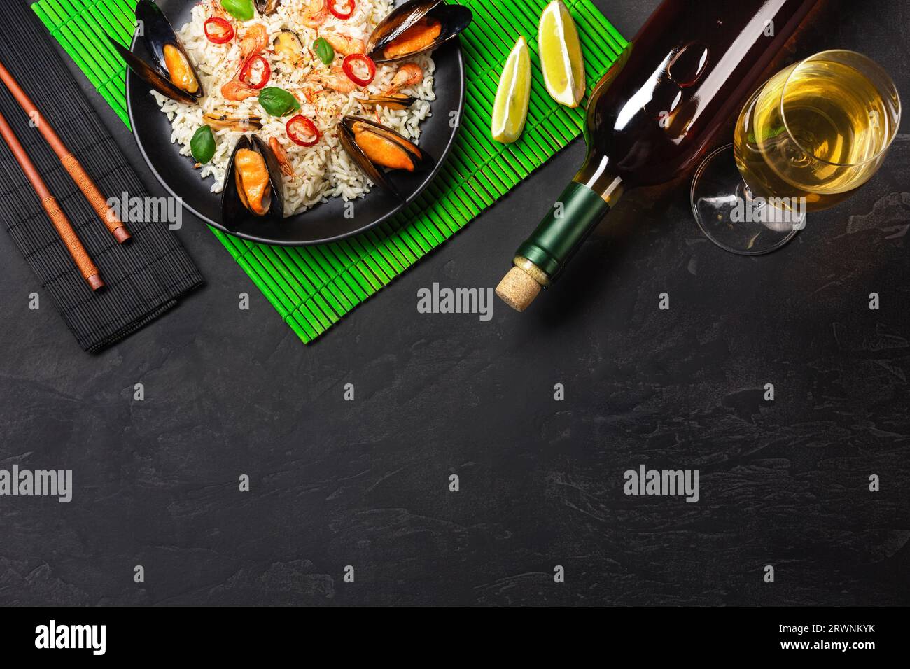 Riso fritto con cozze di pesce, gamberi, basilico in un piatto nero con bottiglia di vino, vetro, limone, bacchette su tappetino di bambù verde e tavolo in pietra. T Foto Stock