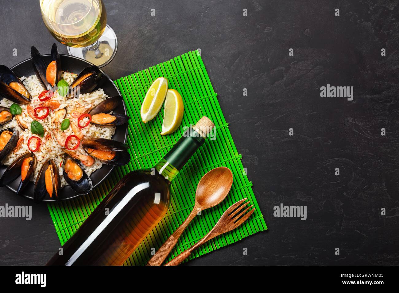 Riso fritto con cozze di pesce, gamberi, basilico in un piatto nero con vetro, limone, cucchiaio di legno e forchetta su tappetino di bambù verde e tavola in pietra. In alto Foto Stock
