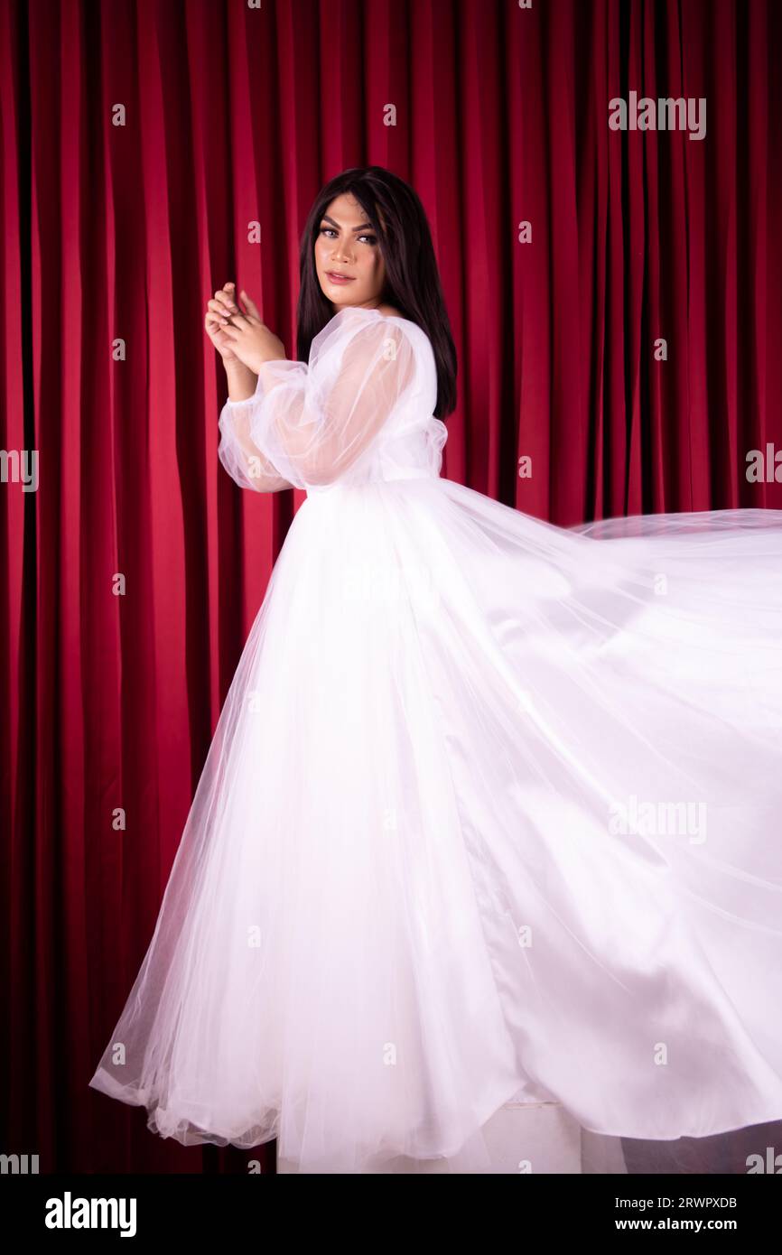 Bella donna asiatica in piedi con una faccia felice in un abito da sposa bianco davanti alla tenda rossa all'interno dello studio Foto Stock