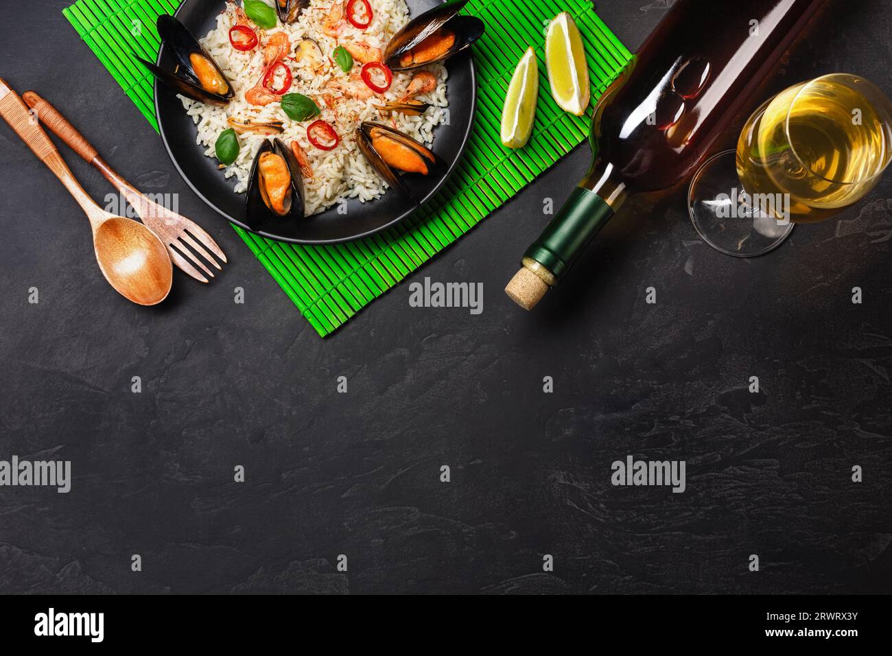 Riso fritto con cozze di pesce, gamberi, basilico in un piatto nero con vetro, limone, cucchiaio di legno e forchetta su tappetino di bambù verde e tavola in pietra. In alto Foto Stock