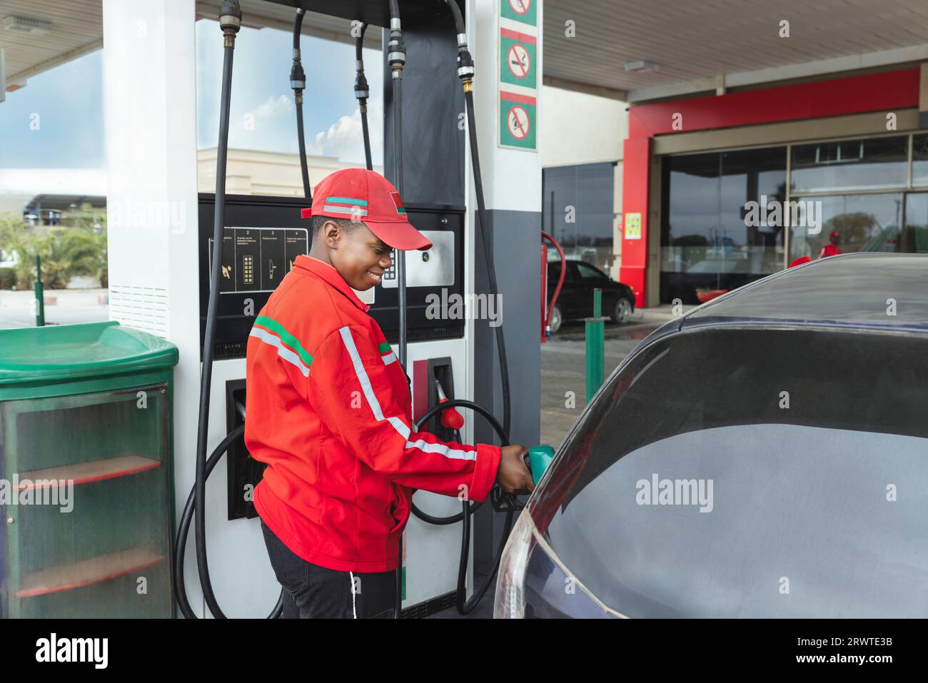 assistente donna afroamericana in uniforme rossa alla pompa del gas, riempiendo con benzina una piccola auto, ugello è nel serbatoio Foto Stock