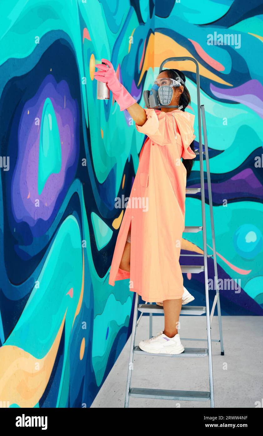 Artista di strada femminile in maschera respiratoria dipinge graffiti colorati su una parete in piedi su una scala. Arte moderna, concetto urbano. Foto Stock