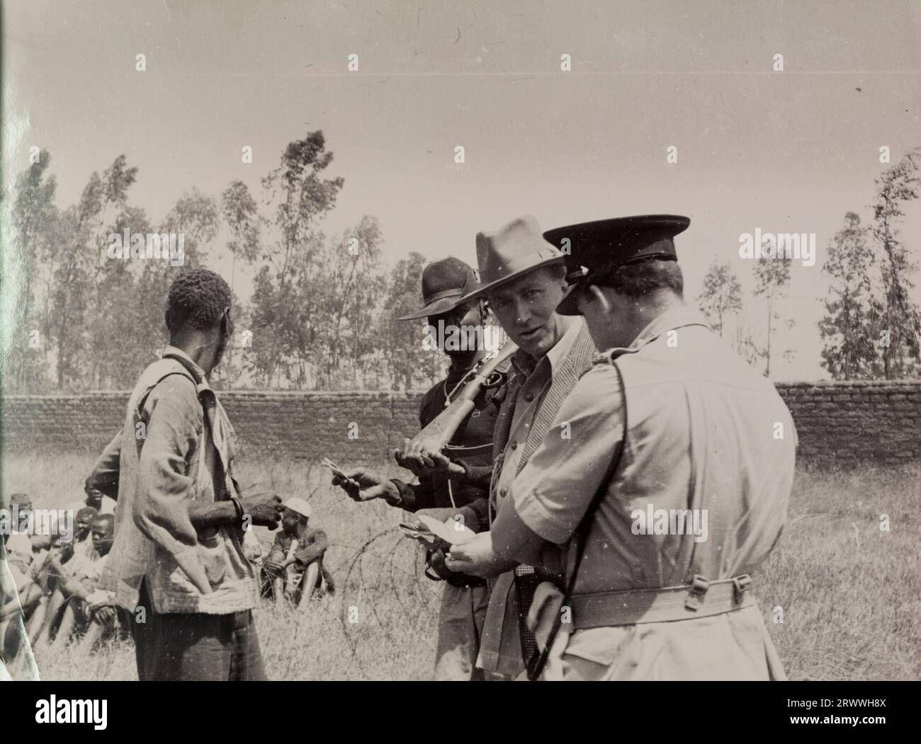 Un contadino e ufficiali militari mettono in discussione un operaio agricolo di Kikuyu. Dietro di lui più Kikuyu siedono a terra circondati da bobine di filo spinato. Didascalia originale: RETURN TO E. HUXLEY, WOOODFOLDS, OAKSEY, MALMESBURY, ENGLAND-The War Against Mau Mau in Kenya: Police raid in una fattoria per i sospettati Mau Mau e interrogare la forza lavoro e i loro amici. Foto Stock