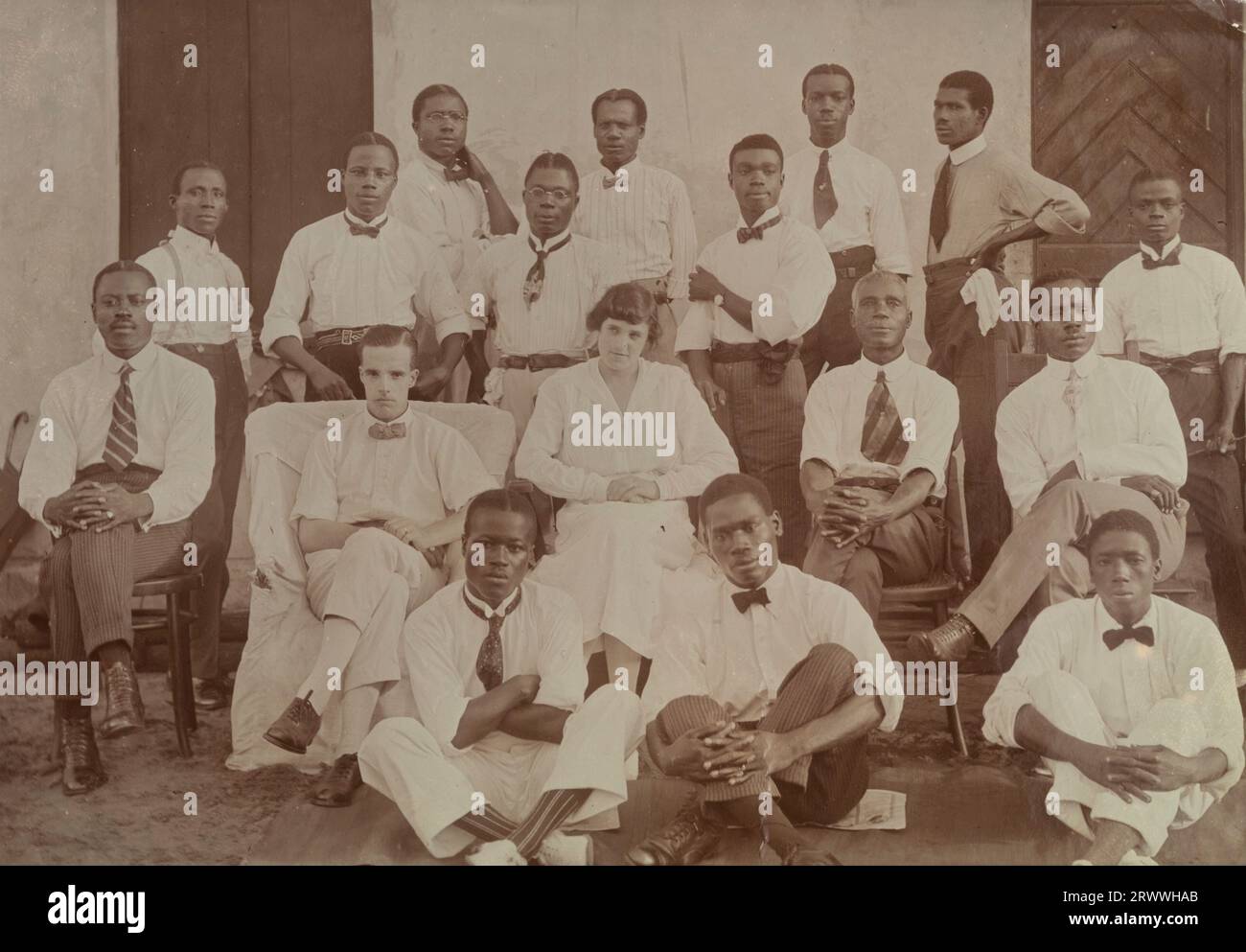 Foto di gruppo del personale d'ufficio presumibilmente della African and Eastern Trade Corporation, costituita nel 1919 dalla fusione di Swanzy's, Millers Ltd e l'African Association. 15 uomini africani in abito da ufficio si mettono in posa con i signori Tamlin: Si sono tolti i giubbotti e sono raggruppati informalmente intorno ai Tamlin. Didascalia del testo originale: Office staff Half Assinie maggio 1920. Didascalia del manoscritto più tardi: Alfred & Dorothy Tamlin seduto al centro. Foto Stock