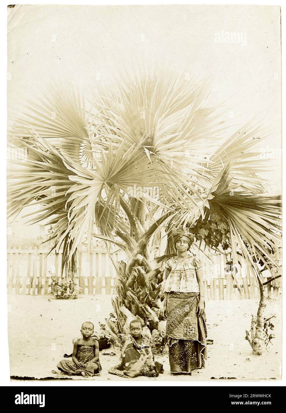 Una donna africana si trova sotto una palma con due bambini seduti a terra accanto a lei. Sono tutti vestiti con coloratissimi capi a motivi geometrici. Foto Stock