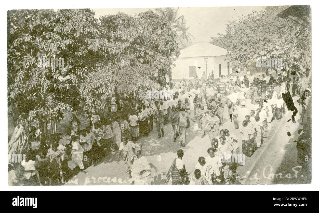 Una strada affollata a Winneba, come una processione del venerdì Santo, attraversa una strada costeggiata da alberi, edifici e spettatori. Due lunghe file di donne vestite in modo intelligente percorrono la strada con una banda che si esibisce al centro di loro e persone che seguono dietro la band che portano bandiere. In negativo: Processione nativa... Gold Coast. Foto Stock