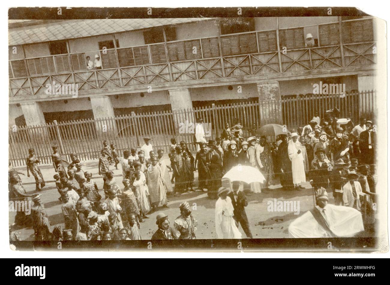 Scena di strada ad Accra nel bel mezzo di un evento in maschera. All'ombra di un grande edificio, una folla si riunisce intorno, molti dei quali con abiti e maschere eleganti. Un gran numero di festaioli sembra prendere in giro gli inglesi, con due coppie vestite in stile occidentale e molte altre in caschi Pith, un cappello e maschere bianche con il naso grande. Un certo numero di spettatori sta guardando lo spettacolo, vestito con tradizionali involucri e copricapi. Didascalia del testo originale: In Accra. Foto Stock