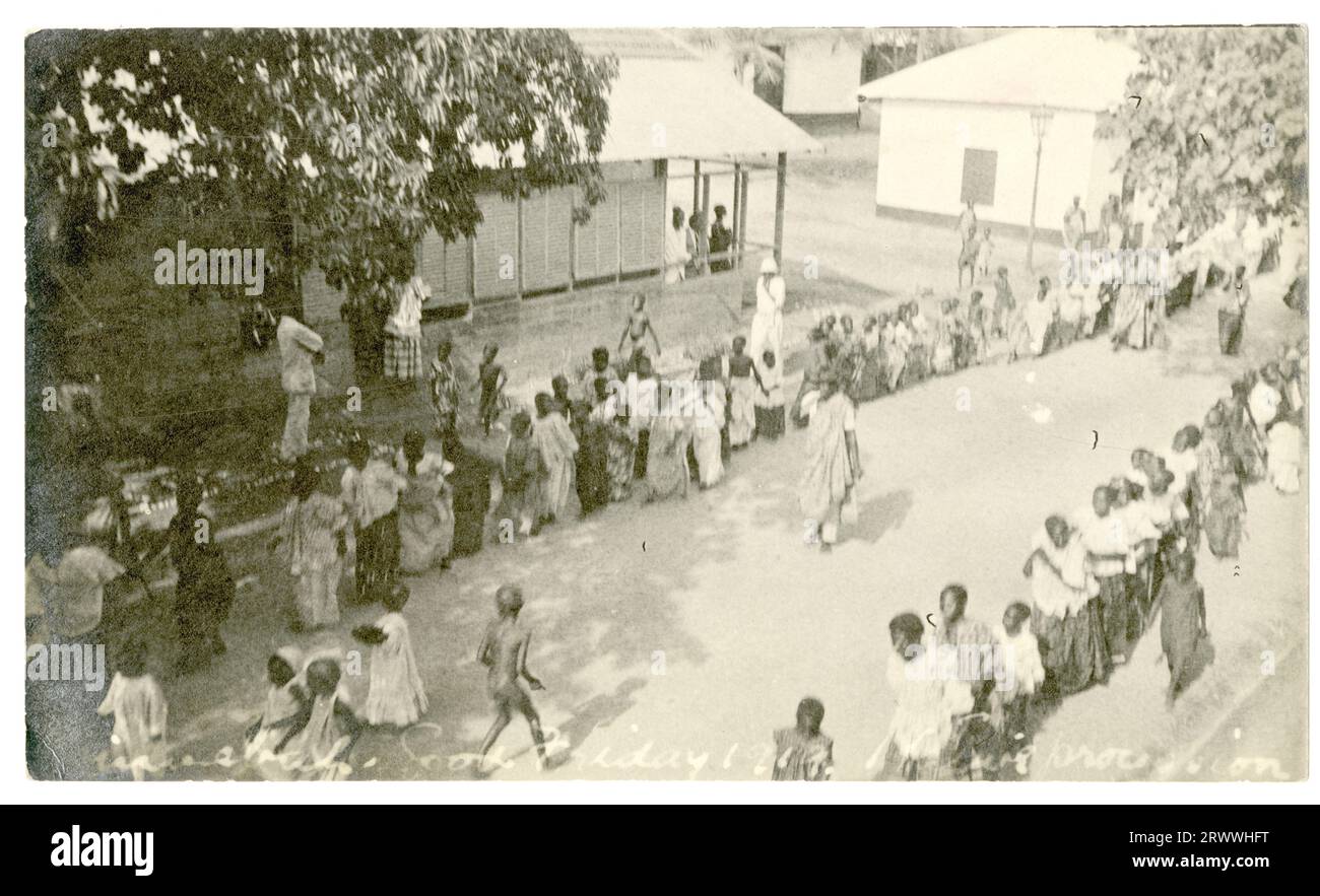 Due lunghe file di donne e bambini attraversano una strada a Winneba, fiancheggiata da alberi ed edifici, come altri guardano. Sono tutti vestiti in modo intelligente. Un piccolo gruppo di bambini con abiti bianchi e cofani cammina in mezzo alla strada tra le file. In negativo: Winnebah. Venerdì Santo 1919. Processione nativa. Foto Stock