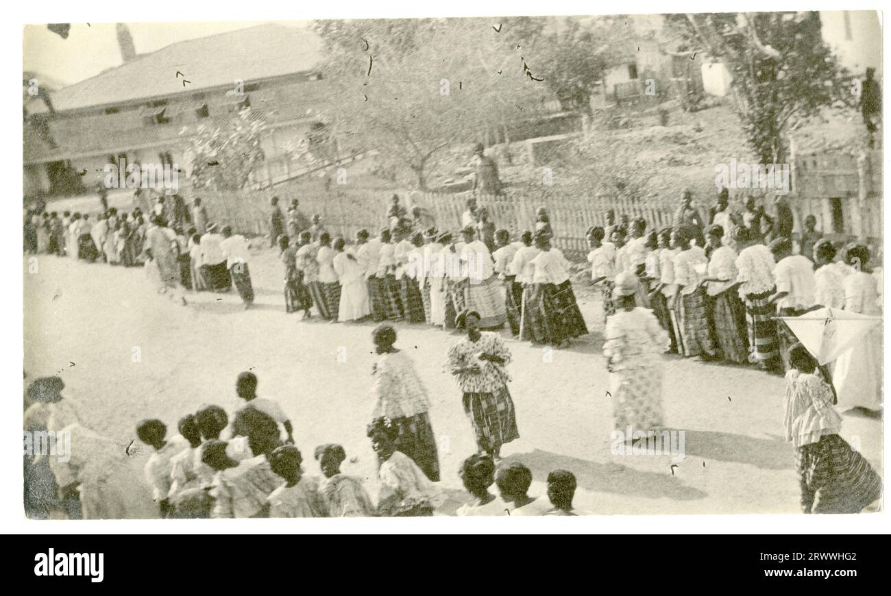 Due lunghe file di donne si radunano in strada, pronte ad iniziare una processione. Tutti i capi sono elegantemente confezionati con camicette bianche e gonne a motivi geometrici. Ci sono edifici visibili dietro di loro. La didascalia di un'immagine abbinata descrive l'evento come una processione del venerdì Santo a Winneba. Foto Stock