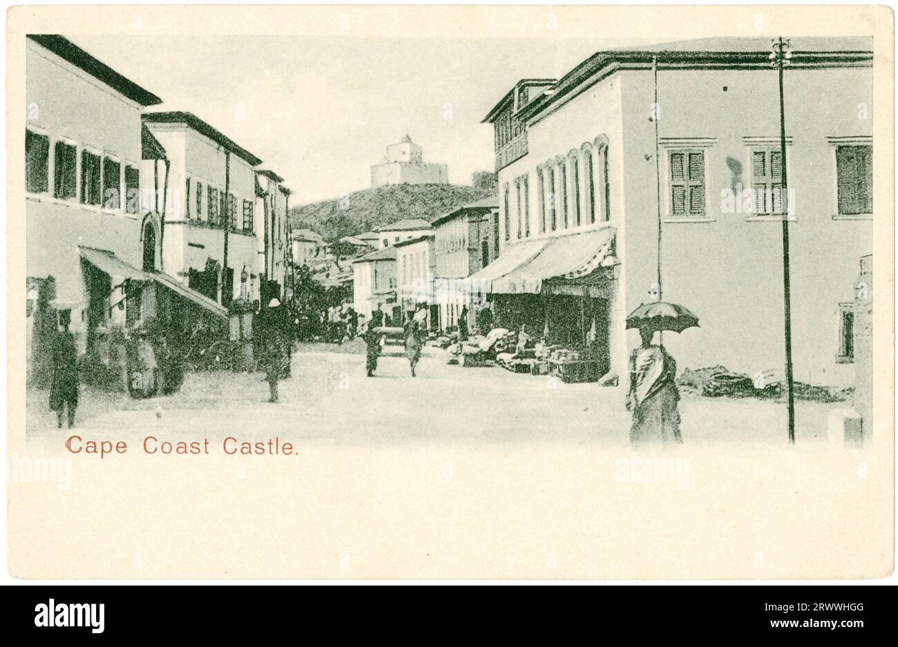 Vista da cartolina del castello di Cape Coast sulla cima di una collina con una strada trafficata sotto. Ci sono negozi su entrambi i lati della strada con ceramiche disposte all'esterno e pedoni africani ed europei. Didascalia stampata: Cape Coast Castle. Foto Stock