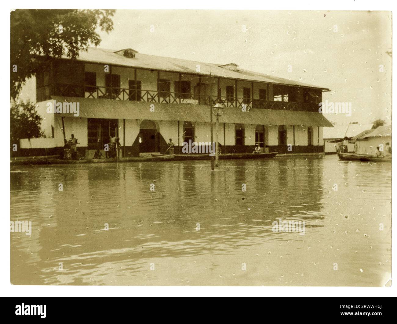 Vista di un grande edificio in stile coloniale circondato dall'acqua, con uomini africani che manovrano punt fino alla porta d'ingresso. Foto Stock