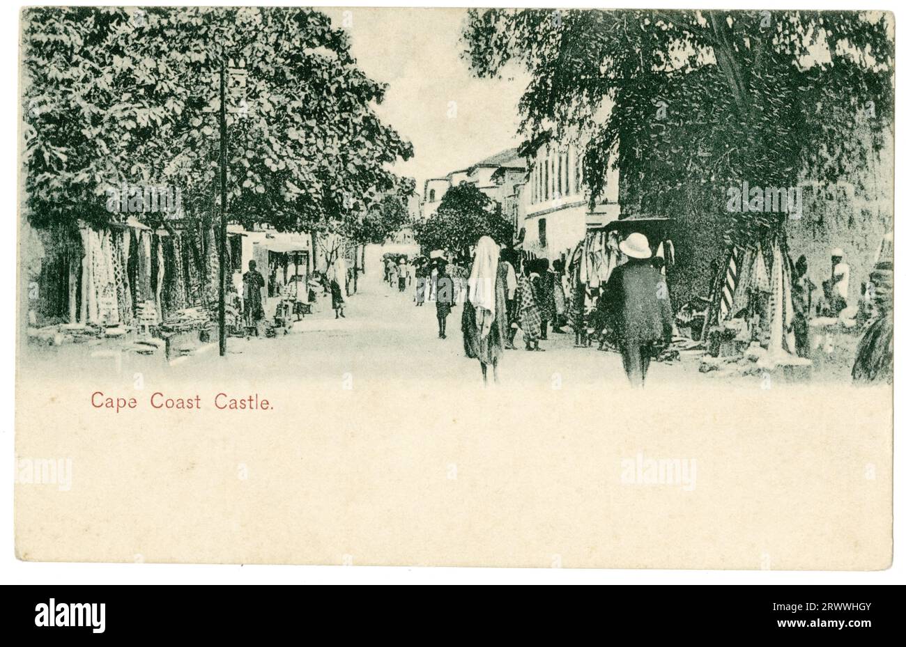Vista da cartolina di una trafficata scena stradale nei dintorni del castello di Cape Coast. Le bancarelle del mercato si trovano su entrambi i lati di una strada alberata, affollata sia dai pedoni europei che africani. Didascalia stampata: Cape Coast Castle. Foto Stock