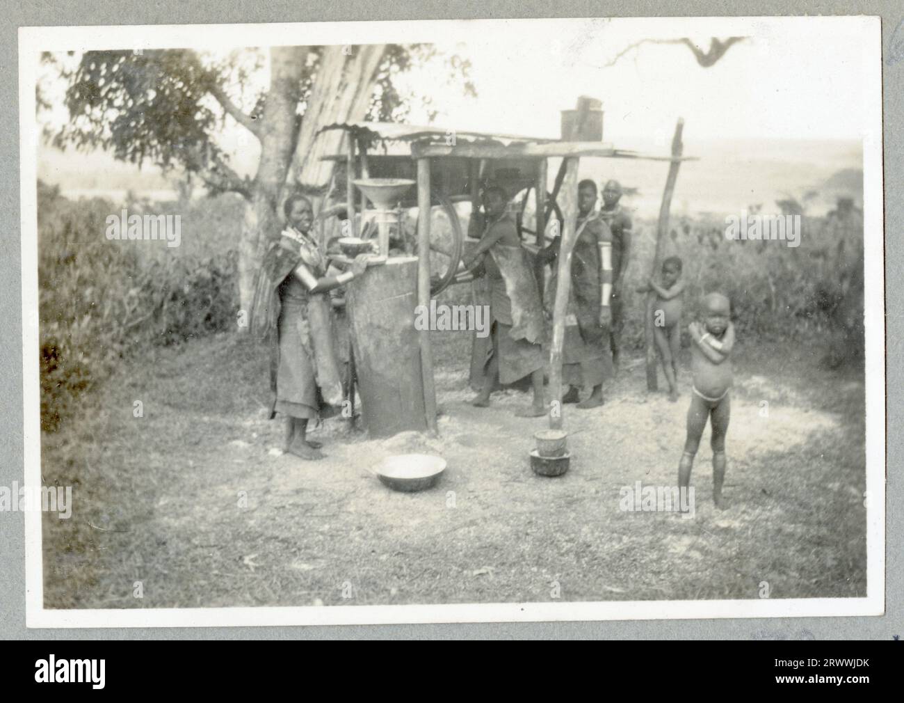 Le donne Nandi stanno sotto un riparo in lamiera di ferro ondulata e girano la ruota di una macchina che macina i cereali per fare la farina. Ci sono anche bambini in piedi. Le donne indossano tutti abiti da pelle e scialli. Didascalia del manoscritto originale: Nandi Women macinazione del mais, località 26 Nandi. Foto Stock