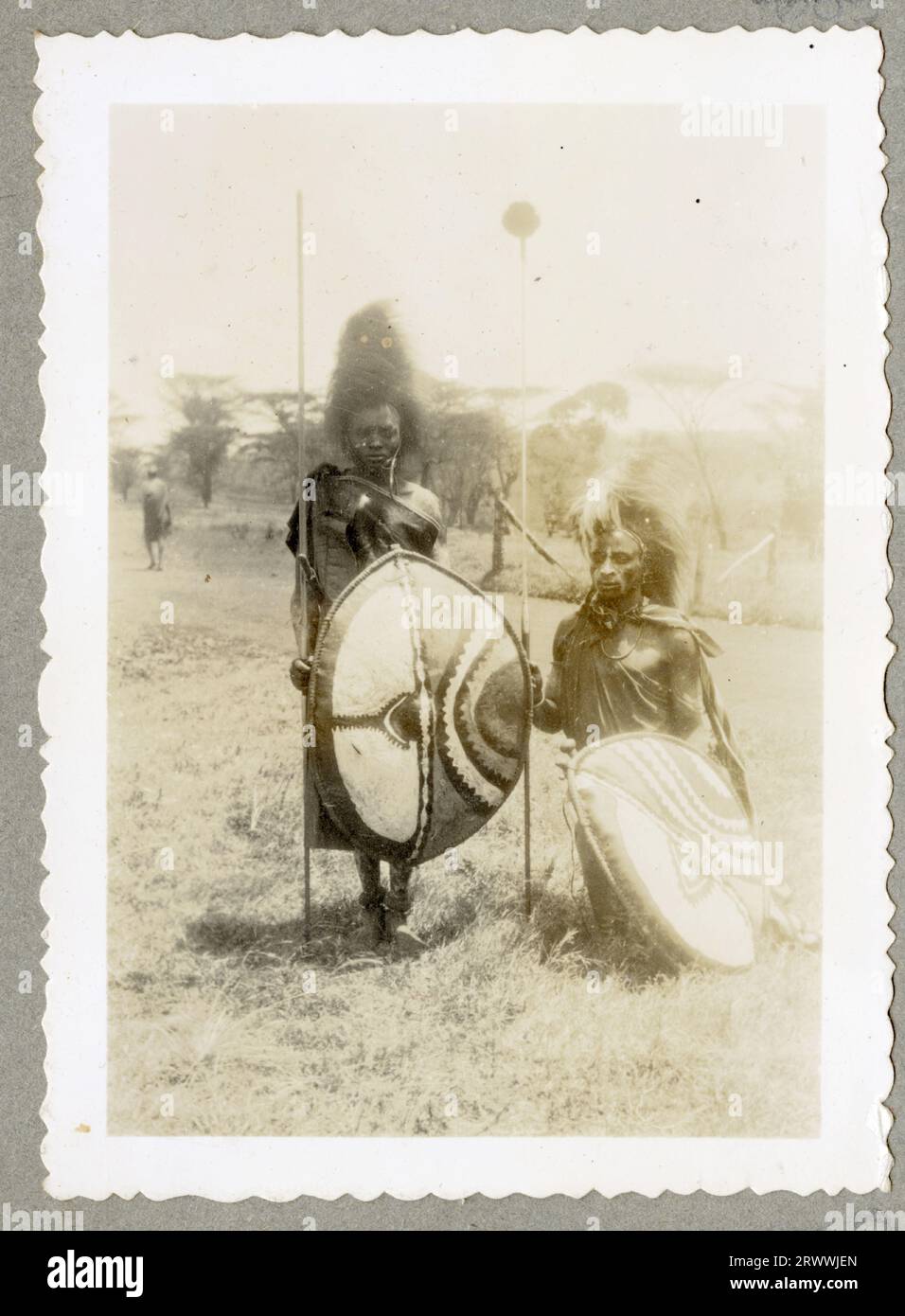 Le donne Nandi stanno sotto un riparo in lamiera di ferro ondulata e girano la ruota di una macchina che macina i cereali per fare la farina. Ci sono anche bambini in piedi. Le donne indossano tutti abiti da pelle e scialli. Didascalia del manoscritto originale: Nandi Women macinazione del mais, località 26 Nandi. Foto Stock