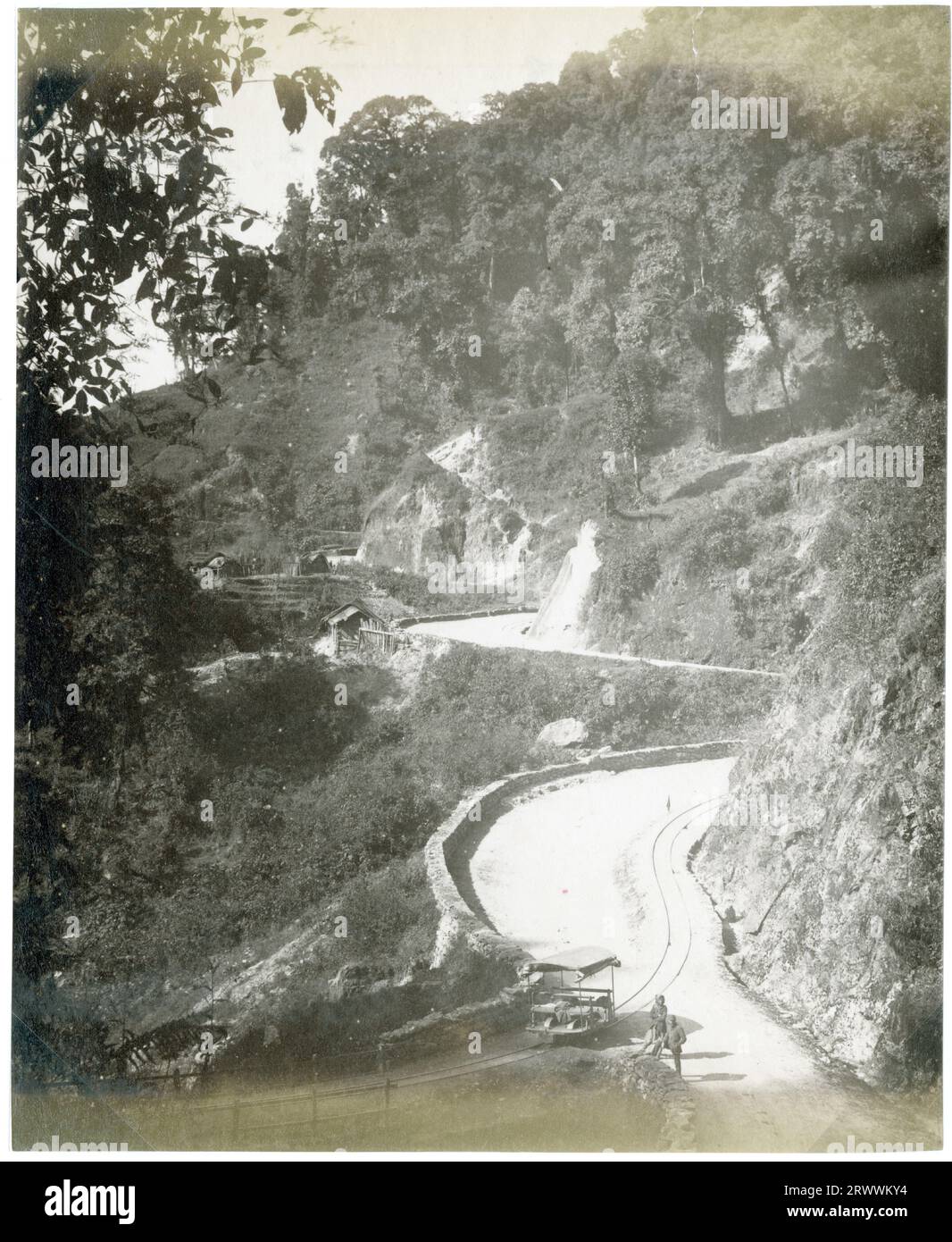Una vista che mostra la curva della ferrovia intorno agli angoli taglienti della collina rocciosa. Ci sono diverse capanne di legno ai lati del binario e due uomini locali in primo piano accanto a un piccolo veicolo di manutenzione ferroviaria. La didascalia recita: Darjeeling H. Ry. Foto Stock