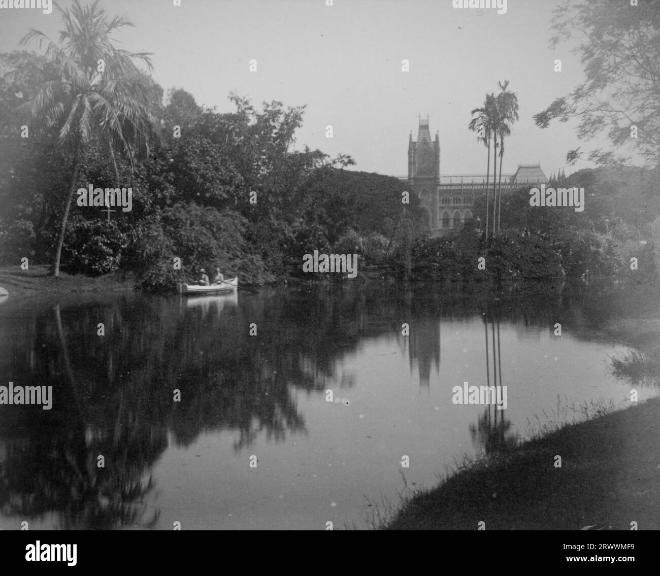 Due uomini europei stanno remando su un fiume a Eden Gardens, arroccati con piante tropicali tra cui palme, con l'alta corte di Calcutta sullo sfondo. Foto Stock