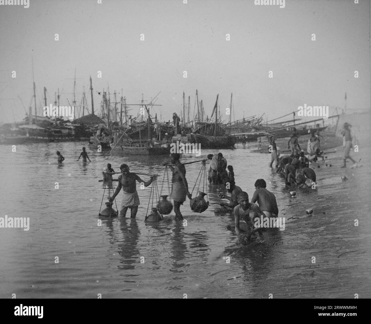 Riva del fiume con sampan e barche da pesca sullo sfondo. Gli uomini indiani possono essere visti lavare o riempire caraffe d'acqua sulle rive e trasportarle sospese dai pali delle spalle. Foto Stock