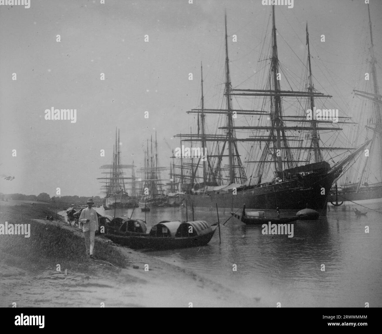 Una fila di barche sampan è ormeggiata su un'insenatura del fiume con grandi navi a vela dietro. Un uomo europeo che indossa un vestito leggero e un casco sta camminando sulla riva del fiume. Foto Stock