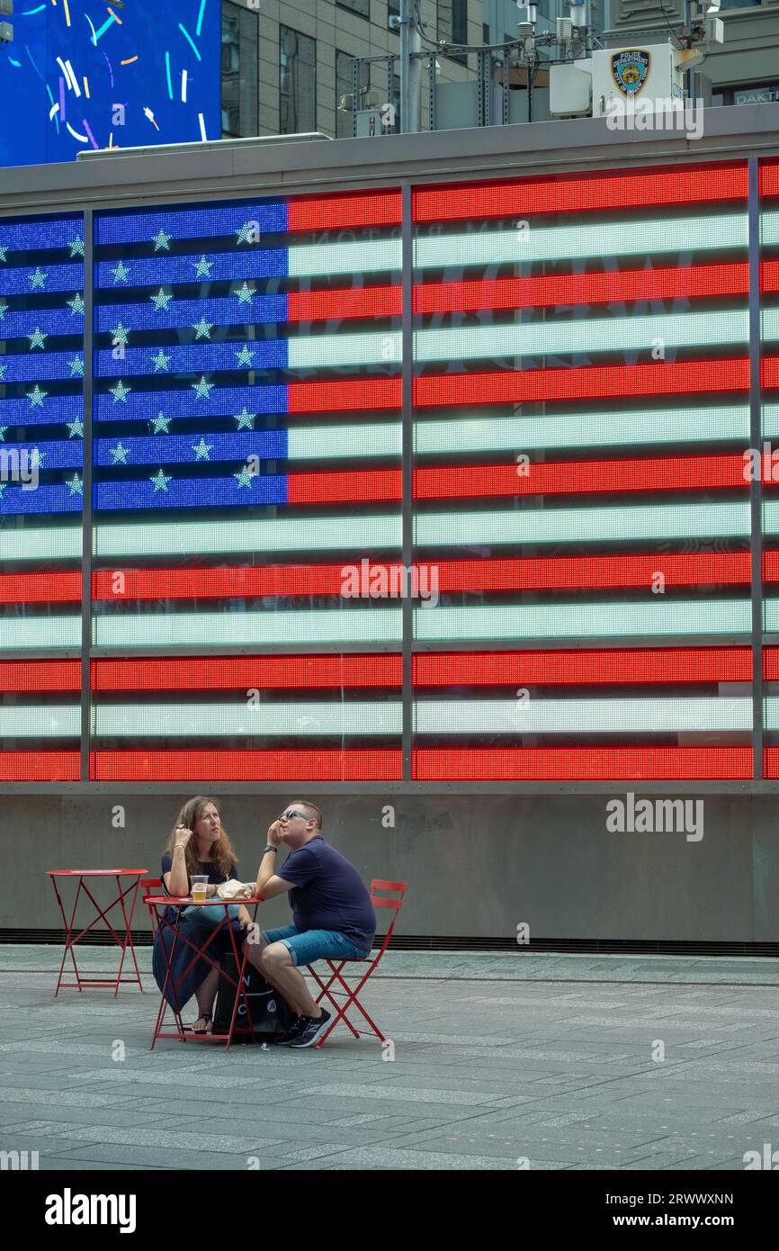 Una coppia di turisti fa una sosta per riposarsi e fare uno spuntino a Times Square di fronte alla gigantesca bandiera americana presso l'ufficio reclutamento dell'esercito degli Stati Uniti. Estate, 2023. Foto Stock