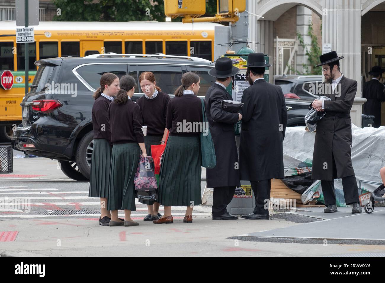 Una scena di strada nella sezione ebraica ortodossa di Williamsburg, Brooklyn, con ragazze in attesa di uno scuolabus e uomini che leggono cartelli. Foto Stock