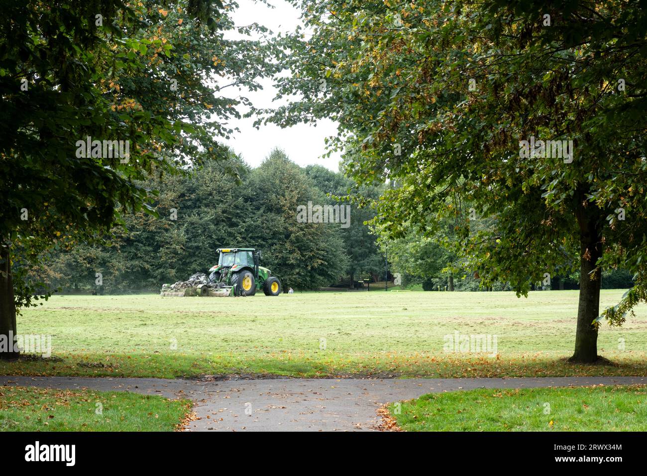 Un appaltatore del consiglio che taglia l'erba in un grande parco pubblico e gioca con un trattore che tira un tagliaerba. Le talee d'erba sono lasciate indietro Foto Stock