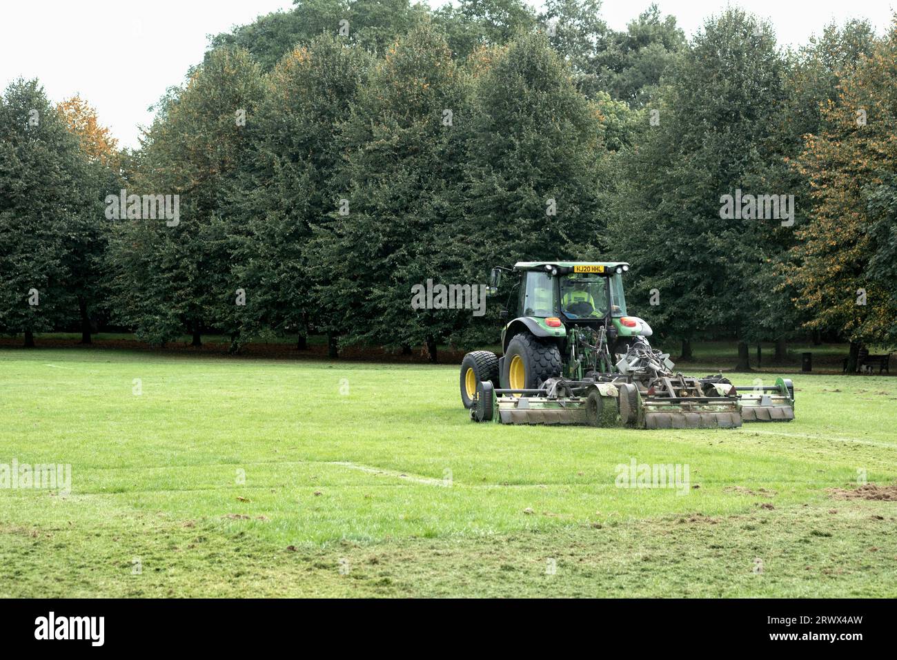 Un appaltatore del consiglio che taglia l'erba in un grande parco pubblico e gioca con un trattore che tira un tagliaerba. Le talee d'erba sono lasciate indietro Foto Stock