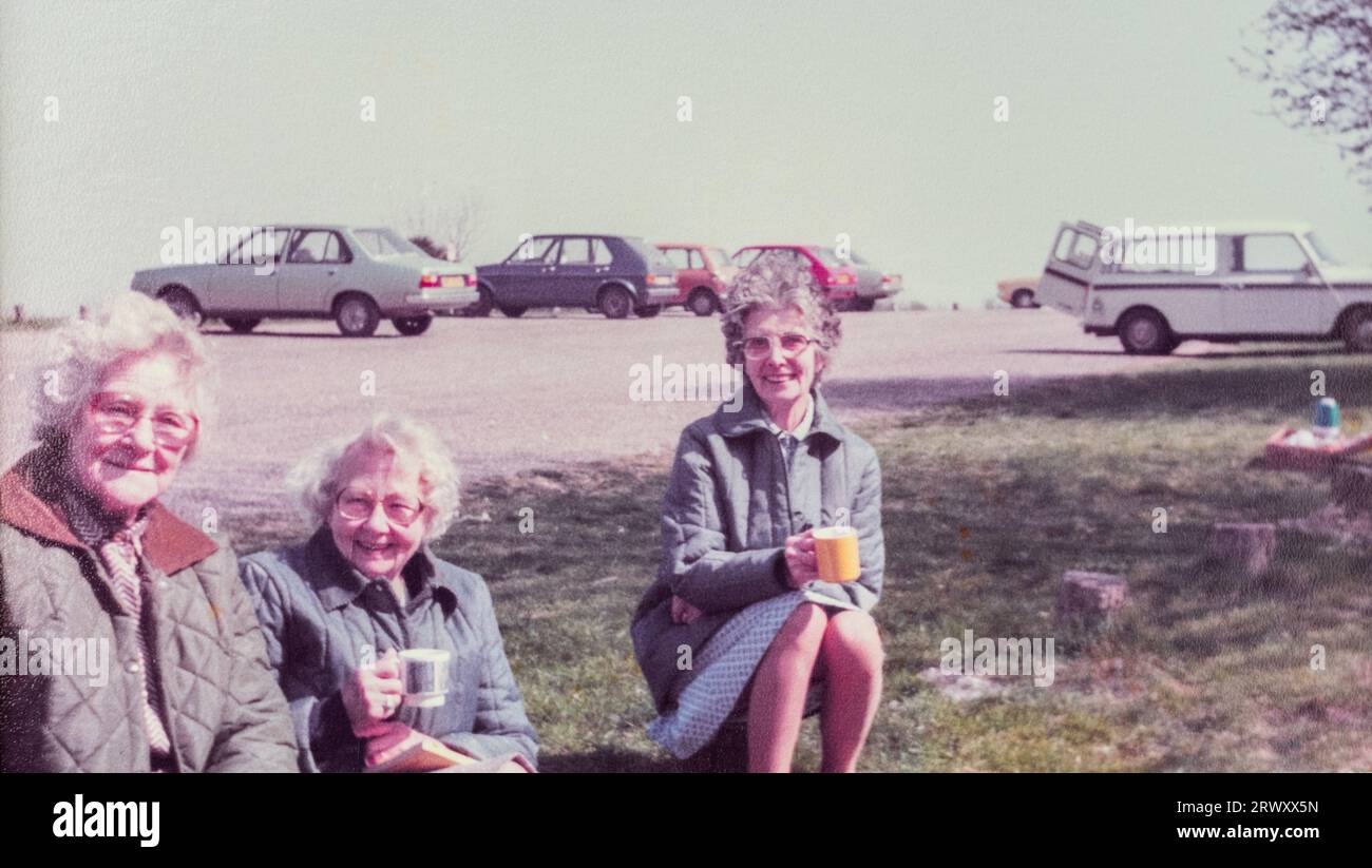 Foto d'archivio di tre donne anziane in un luogo per picnic che bevono una tazza di tè da una fiasca con vecchie auto nel parcheggio sullo sfondo, alla fine degli anni '1970 Foto Stock