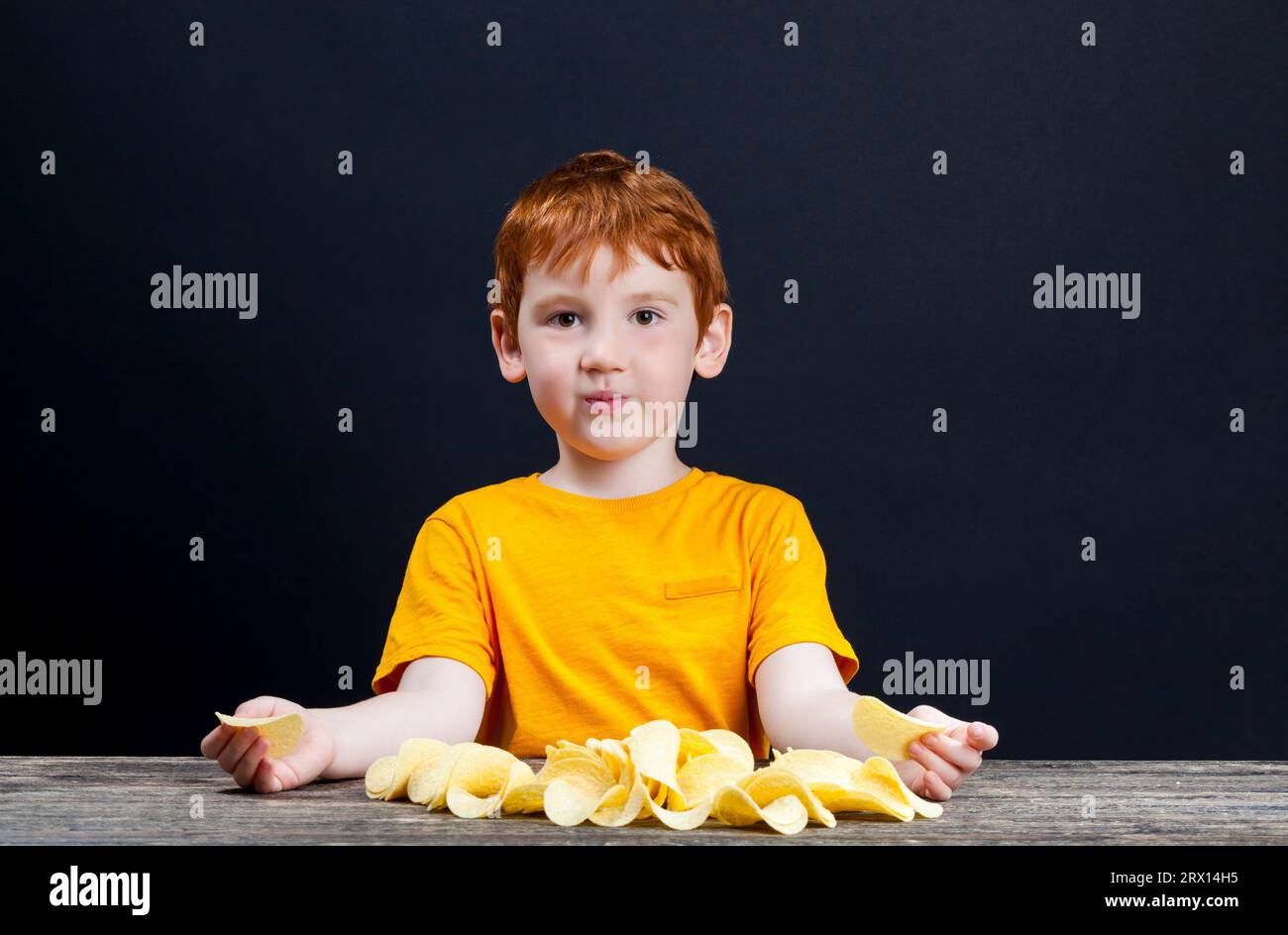 patatine croccanti deliziose che un bambino con i capelli rossi mangia, cibo nocivo, ma che il bambino vuole davvero mangiare, il ragazzo al tavolo, vicino Foto Stock