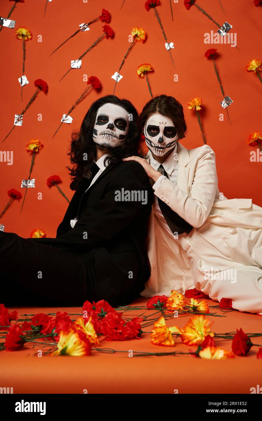 elegante coppia con trucco di teschio di zucchero e abiti seduti sul pavimento in studio rosso con fiori di garofano Foto Stock