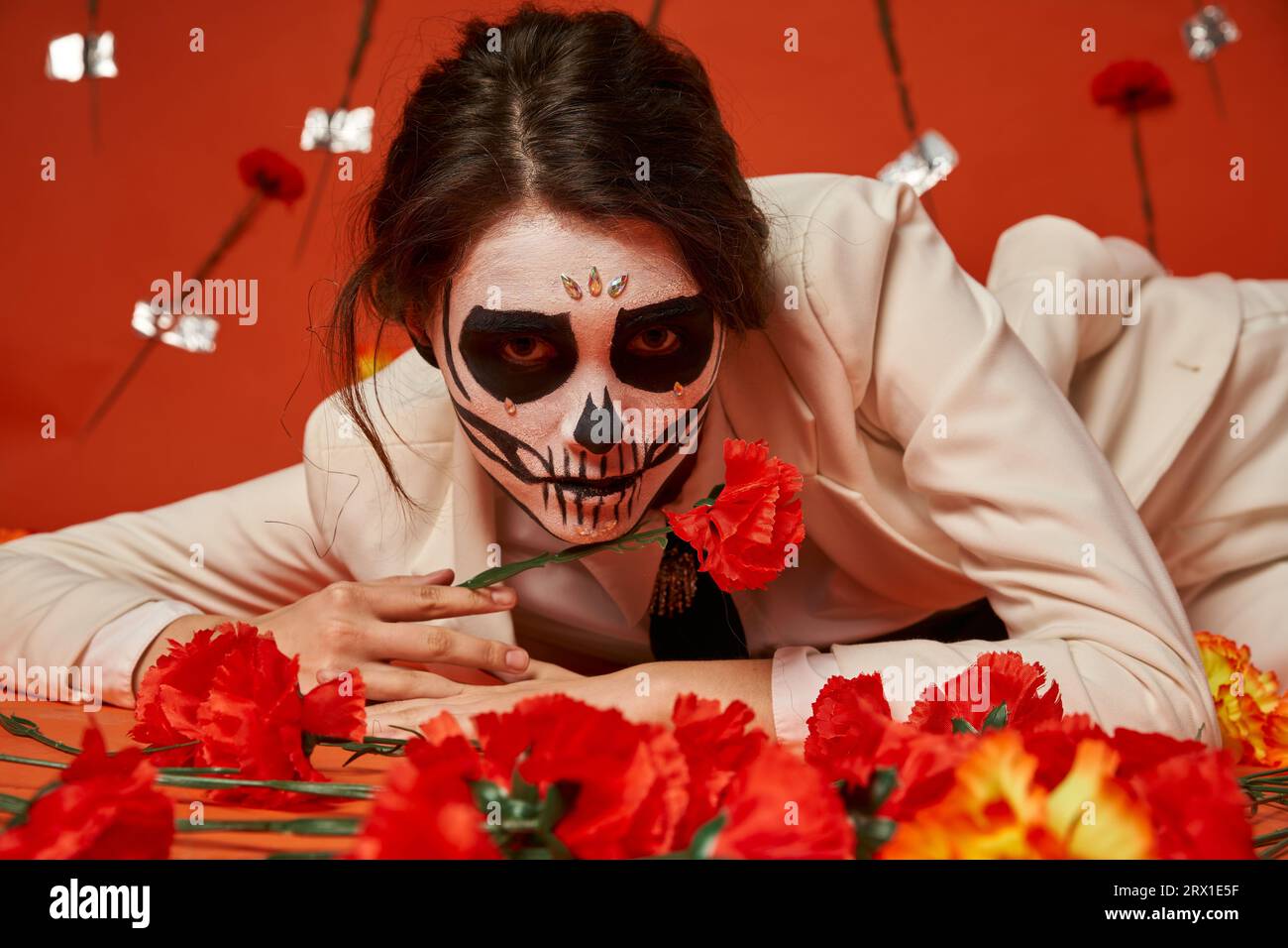 donna elegante con spaventoso trucco catrina sdraiata e guardando la macchina fotografica vicino ai fiori su sfondo rosso Foto Stock