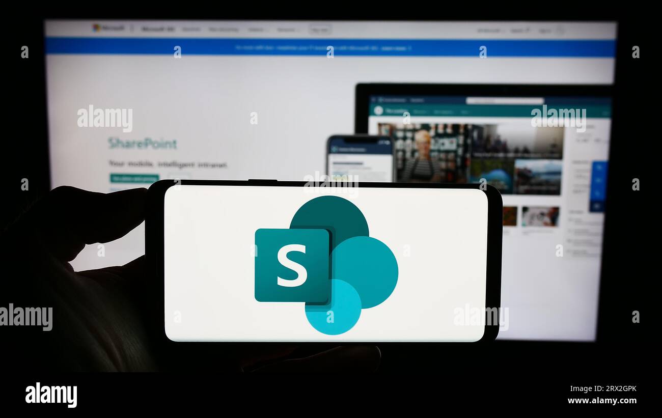 Persona che tiene il cellulare con il logo della piattaforma collaborativa Microsoft SharePoint sullo schermo davanti alla pagina Web dell'azienda. Concentrarsi sul display del telefono. Foto Stock
