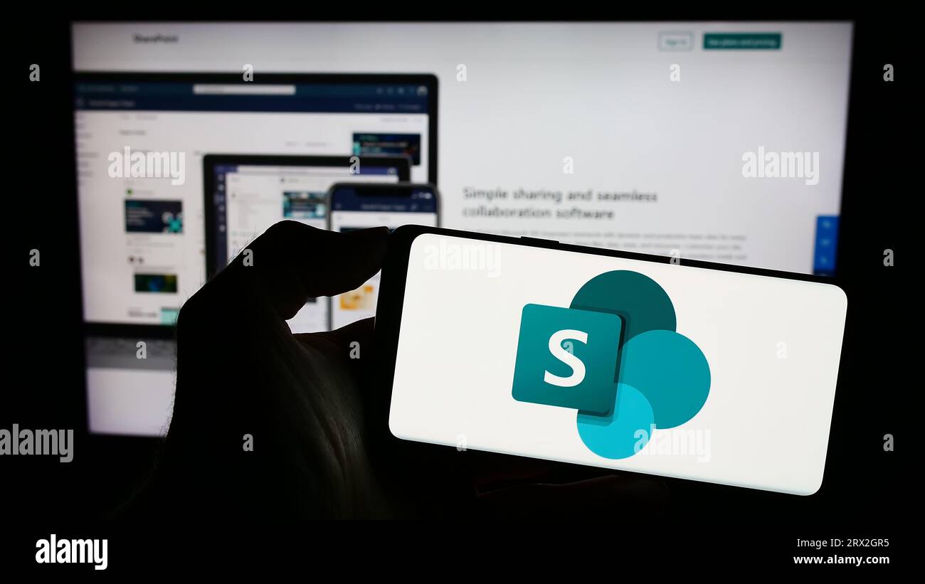 Persona che possiede uno smartphone con il logo della piattaforma collaborativa Microsoft SharePoint sullo schermo di fronte al sito Web. Concentrarsi sul display del telefono. Foto Stock