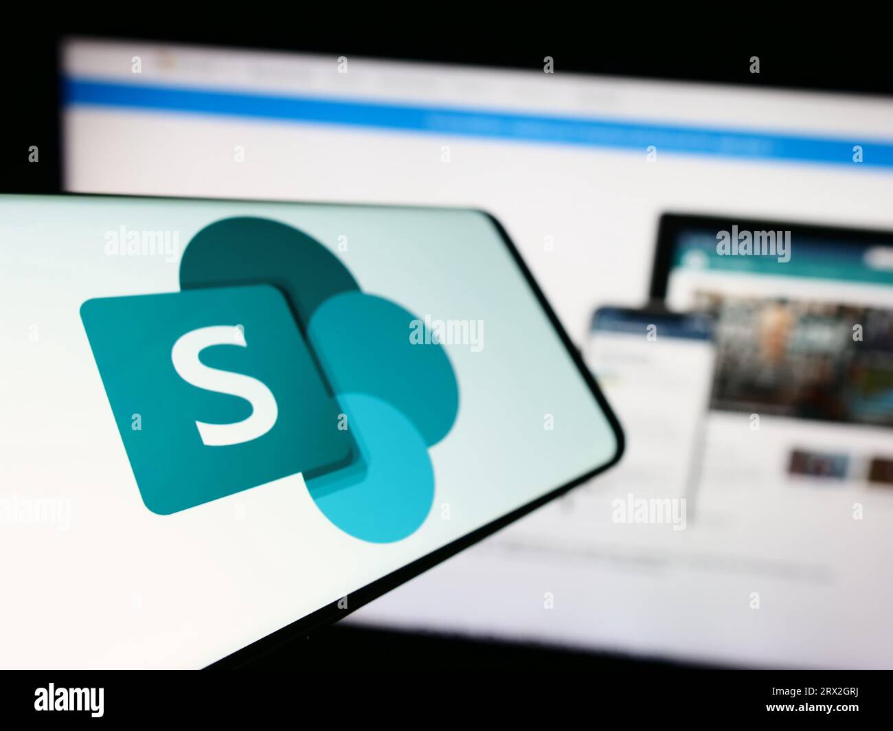 Cellulare con logo della piattaforma collaborativa Microsoft SharePoint sullo schermo di fronte al sito Web aziendale. Mettere a fuoco sul lato sinistro del display del telefono. Foto Stock
