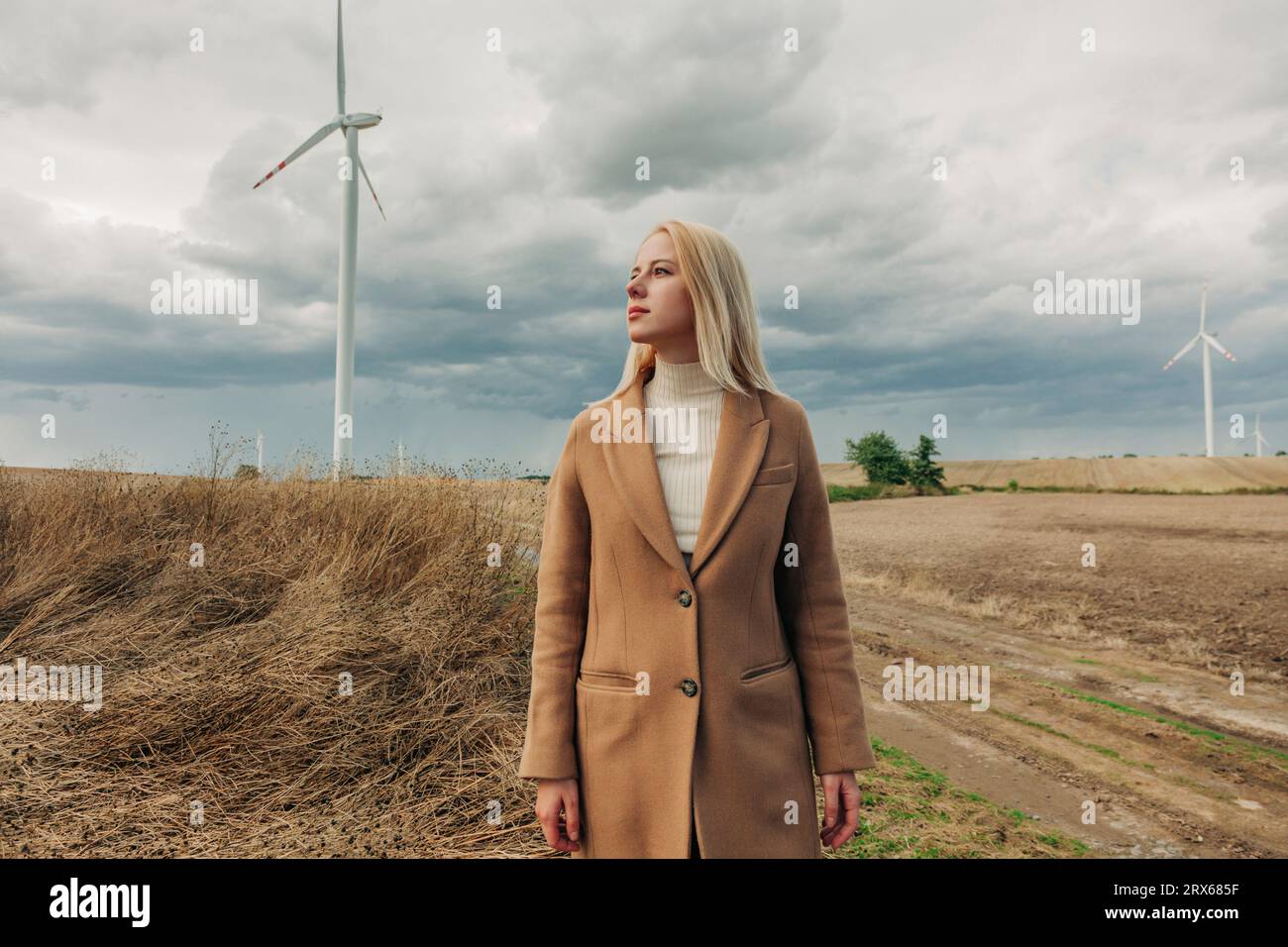 Donna bionda in piedi davanti alla turbina eolica sotto il cielo nuvoloso Foto Stock