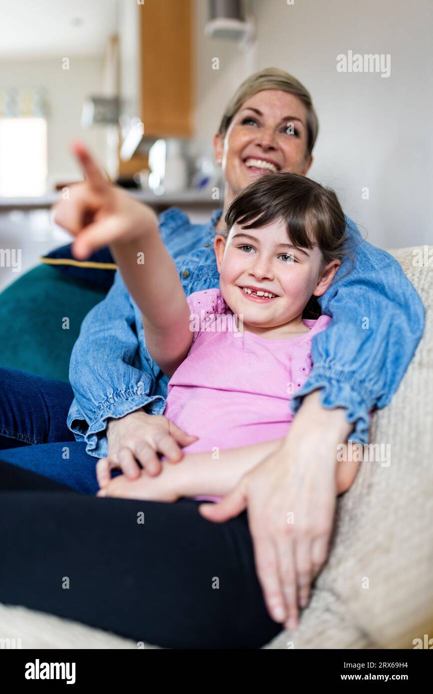 Ragazza sorridente che punta e siede accanto alla madre in salotto Foto Stock