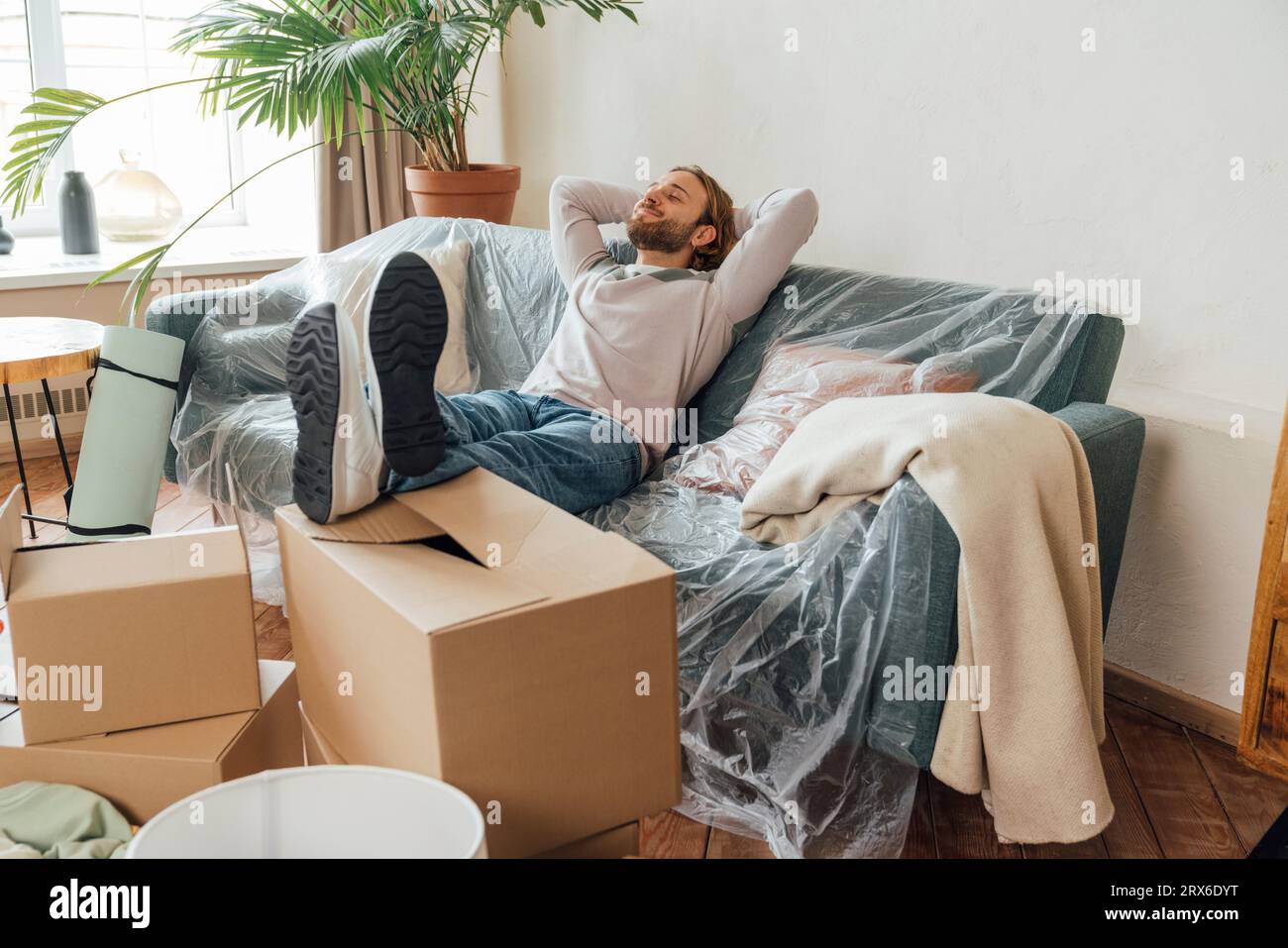 Uomo sorridente con le mani dietro la testa seduto sul divano a casa Foto Stock