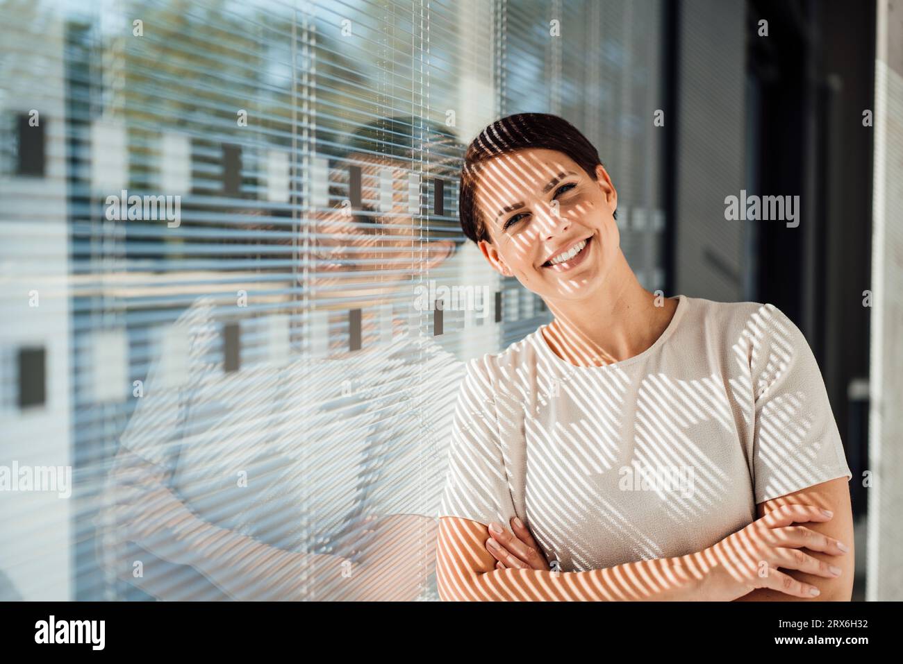 Donna d'affari sorridente con le braccia incrociate appoggiate alla finestra Foto Stock